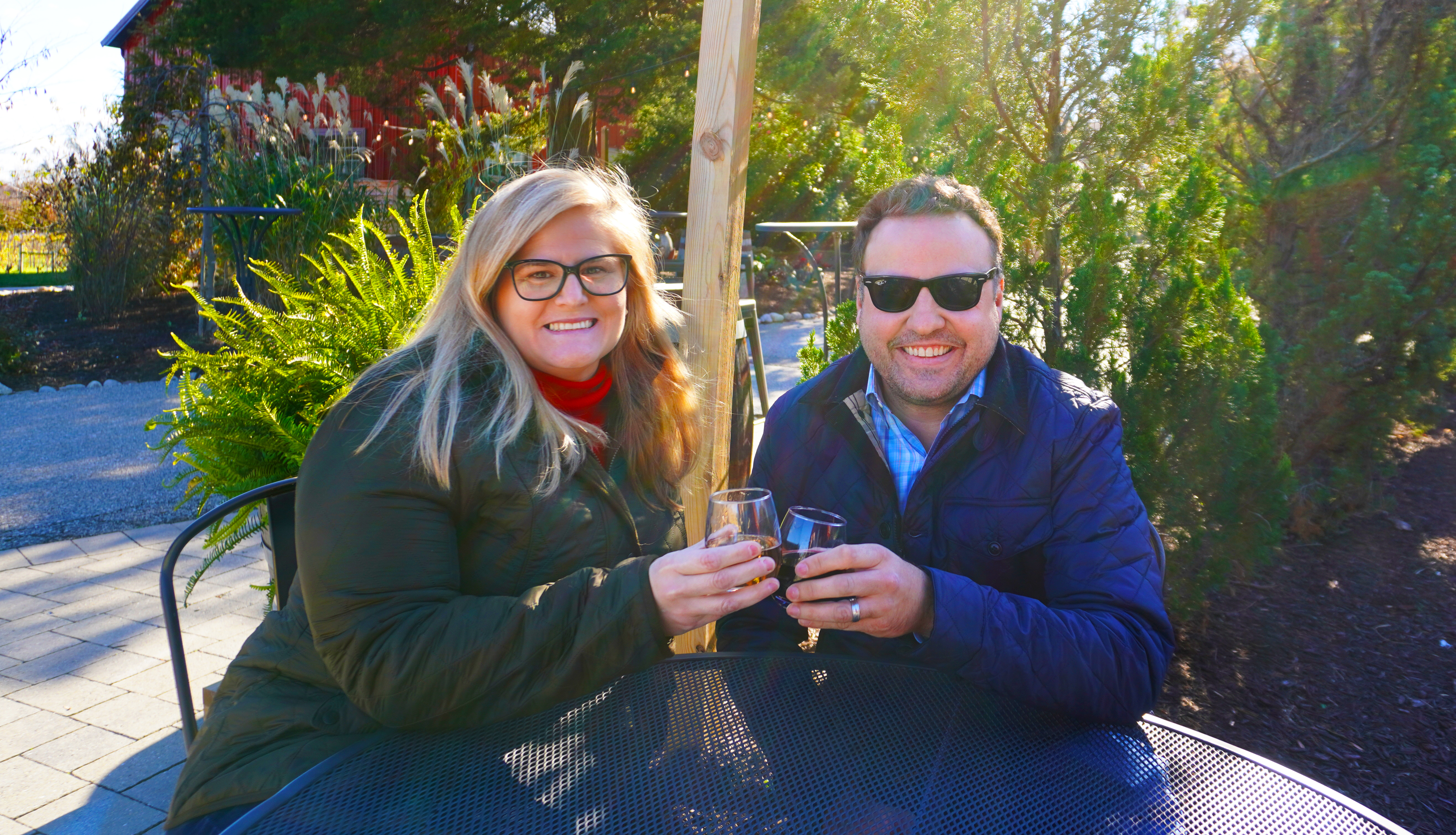 A man and woman sitting at a table outdoors holding glasses of wine.