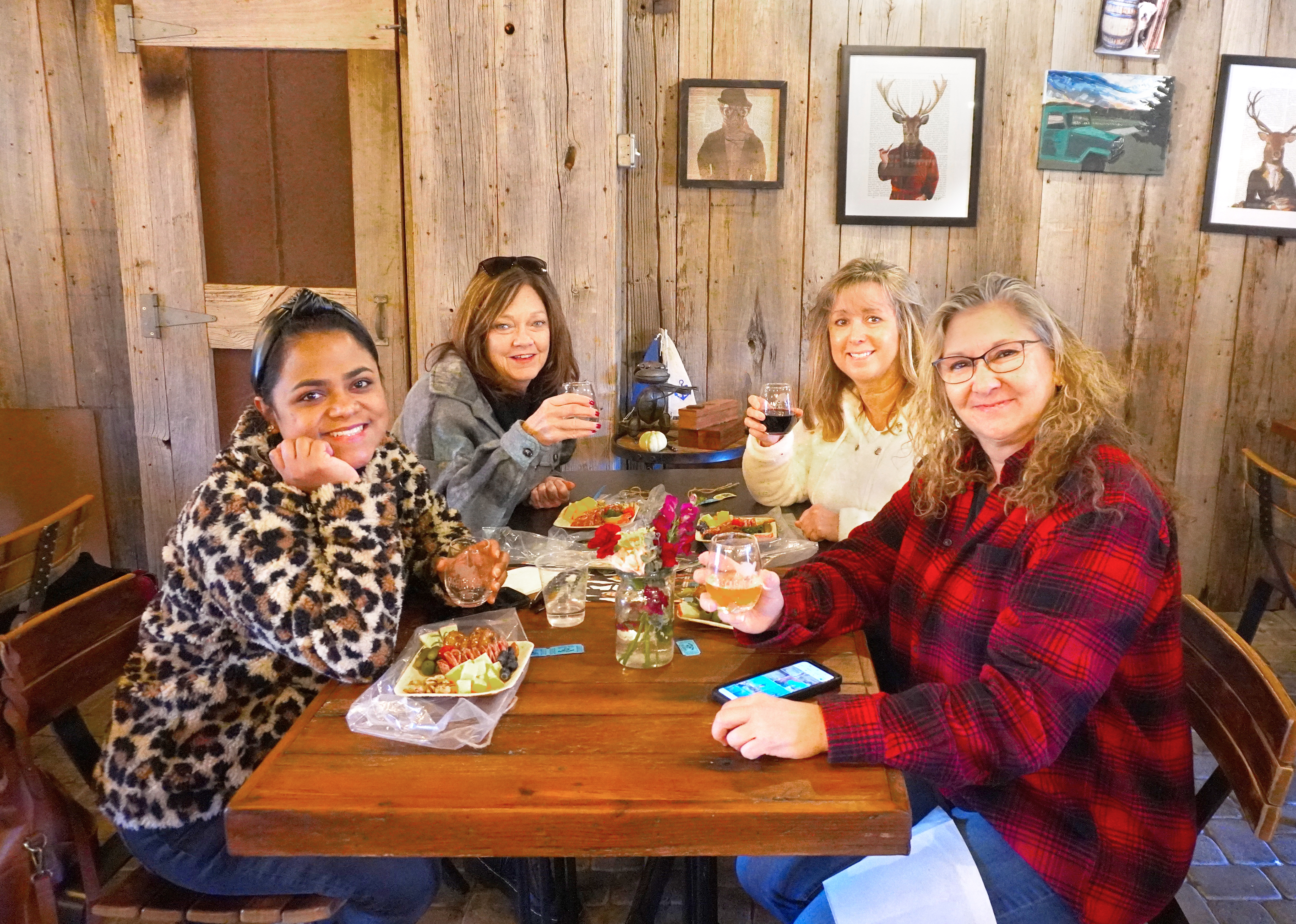 Four smiling women sitting at a table indoors with food and glasses of wine.