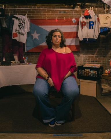 A woman sits in front of a Puerto Rican flag and other cultural items.