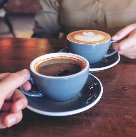 Two mugs of coffee on a table, the handle of each mug is being held.