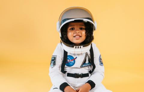 Female child with dark curly hair wearing an astronaut costume in front of a yellow background