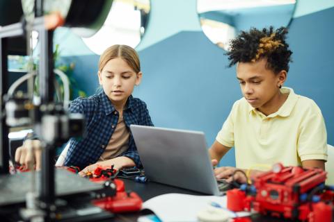 Male child in yellow shirt works on laptop next female child in blue checkered shirt.