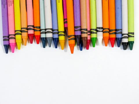 the image shows a row of crayons against a white background. Many of the crayons colors are yelow, green, blue, red, orange, purple, and pink. 