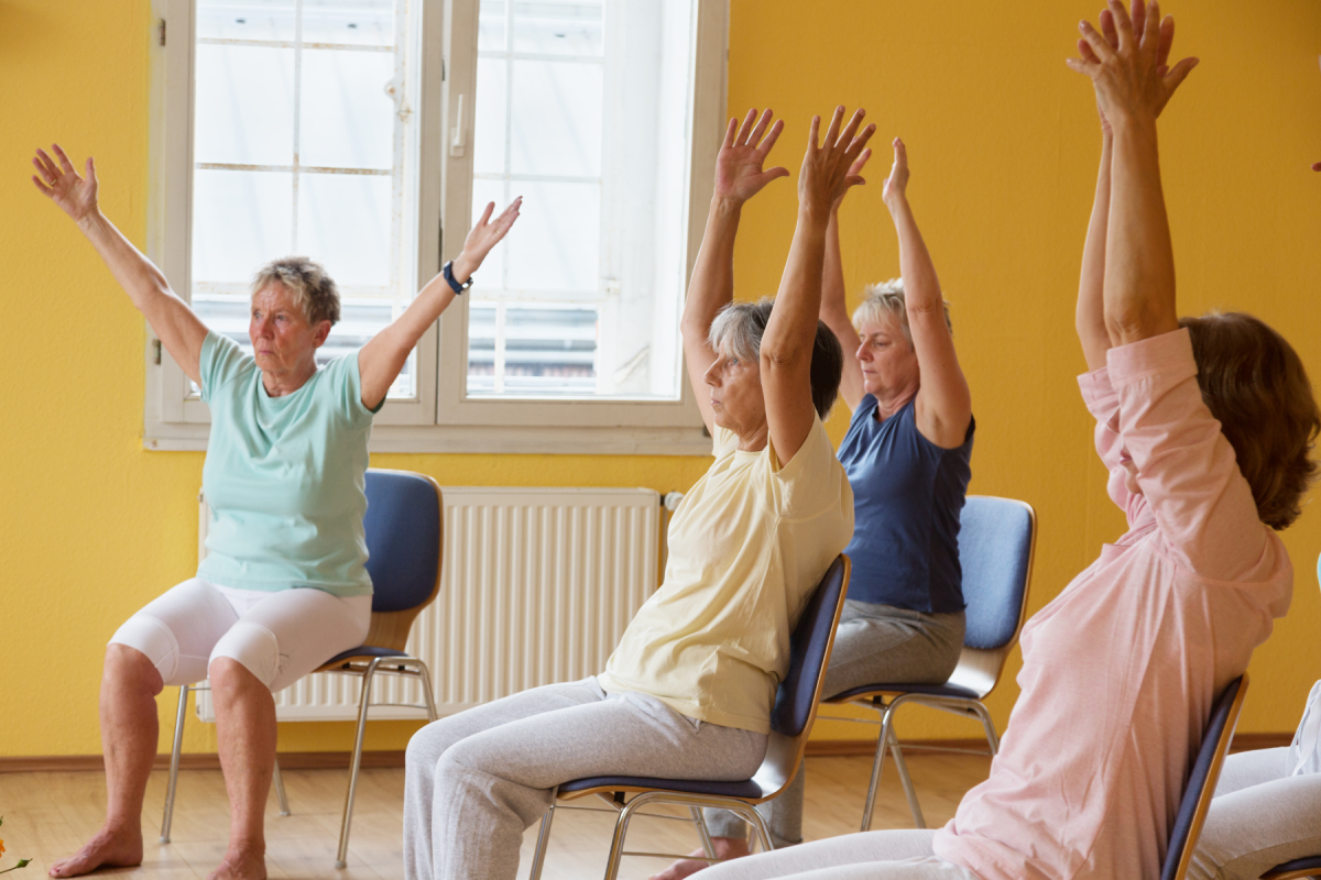 Group chair yoga class, four people sitting in chairs, stretching their arms over their heads