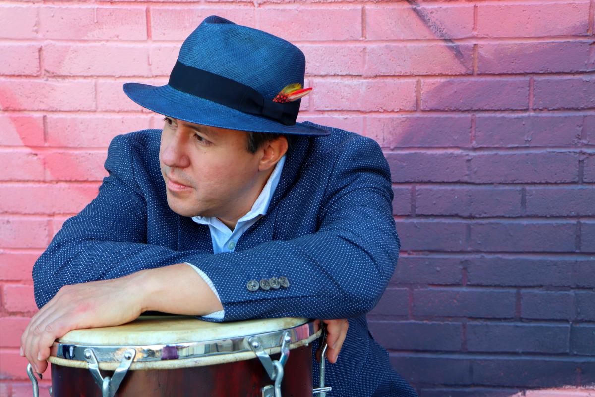 A musician with a blue hat and blue blazer, hands resting on drum