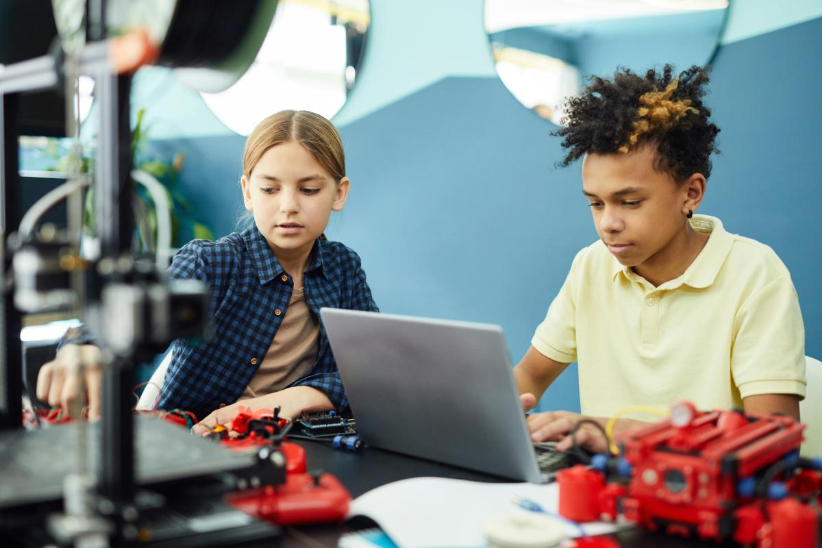 Male child in yellow shirt works on laptop next female child in blue checkered shirt.