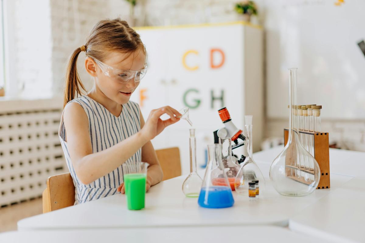 Female child wearing striped dress and safety goggles holds a pipette over a beaker of blue liquid, surrounded by beakers and test tubes.