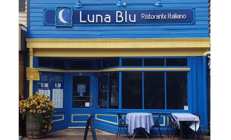 A blue storefront with the sign "Luna Blu Ristorante Italiano" above the entrance. There are two small tables with white tablecloths and blue chairs outside, along with a potted plant with yellow flowers near the entrance.