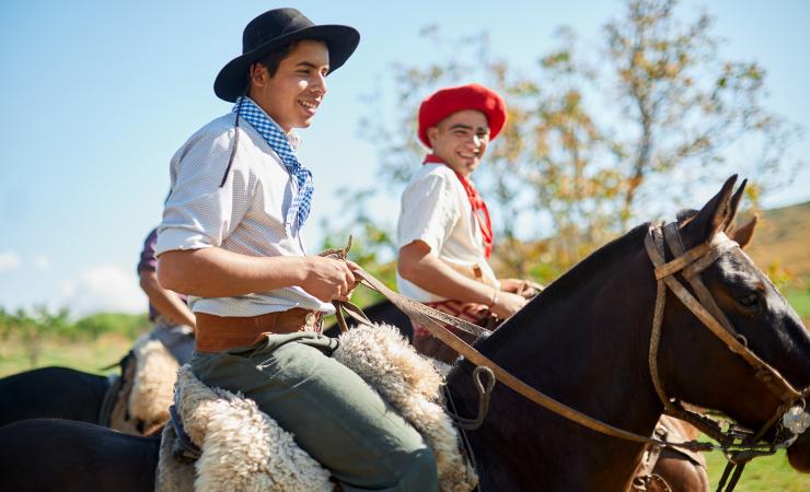 Two people, wearing traditional hats and scarves, are riding horses on a sunny day. One wears a black hat and blue scarf; the other wears a red hat and red scarf.